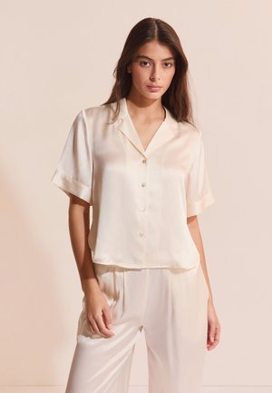 Kobieta z długimi ciemnymi włosami, ubrana w kremową koszulę z krótkim rękawem na guziki oraz dopasowane, luźne spodnie, na beżowym tle.