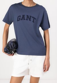 Navyblauwe katoenen t-shirt met korte mouwen en een ronde hals, met een groot "GANT" logo op de borst. Gecombineerd met witte shorts.