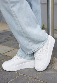 Białe trampki z teksturowaną skórzaną powierzchnią, perforacjami na czubku, grubą białą podeszwą oraz charakterystycznym akcentem po boku. Noszone z jasnoniebieskimi jeansami.