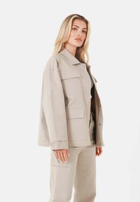 Beige oversized jacka med krage, stora fickor och tryckknappar. Matchas med byxor i samma färg, som har en avslappnad passform och slät textur.