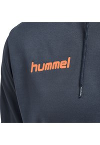 Sweat à capuche bleu marine en matériau texturé, avec un logo "hummel" orange sur le devant et une étiquette de marque sur la couture latérale.