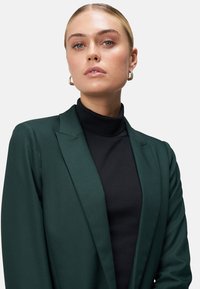 Groene blazer met gestructureerde revers, gecombineerd met een zwarte coltrui. Glad stoffen textuur, minimalistisch design en zichtbare oorbellen met hoop.