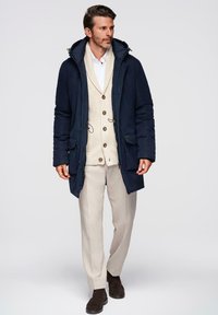 Marine parka met een capuchon, voorzien van een ritssluiting en trekkoorden. Gelaagd over een lichtbeige gebreide cardigan en beige broek. Bruine laarzen.