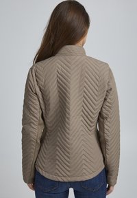 Jasnobrązowa pikowana kurtka z wzorem w jodełkę, wysokim kołnierzem i dopasowanym krojem, zestawiona z ciemnoniebieskimi jeansami.