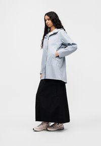 Svetlomodrá nepremokavá bunda s kapucňou, bočnými vreckami a voľným strihom, kombinovaná s čiernou maxi sukňou a svetlými športovými topánkami.