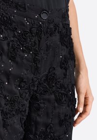 Črne hlače z teksturirano tkanino, okrašene z aplikacijami iz tkaninskih vrtnic in bleščicami. V pasu je okrogla gumb.