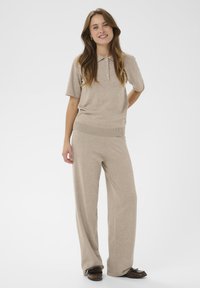 Haut polo beige en maille avec manches courtes et trois boutons, associé à un pantalon à jambes larges assorti. Les deux vêtements ont une texture douce.