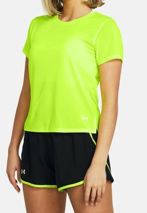 Vrouw die een fel neon groene sportshirt met korte mouwen en zwarte korte broek met groene bies draagt, staand tegen een witte achtergrond.
