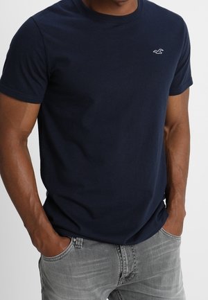 Mand iført en ensfarvet marineblå t-shirt med korte ærmer med lille hvid fuglelogo på brystet og lysegrå jeans med hænderne i lommerne.
