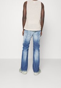 Camiseta de tirantes beige con canalé, vaqueros acampanados azules con desvanecidos y ligeras arrugas, y zapatillas grises, fotografiados desde atrás.