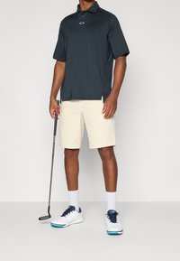 Polo noir à manches courtes avec logo, shorts crème, chaussures blanches avec accents bleus, et un club de golf à la main.