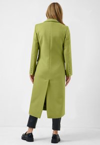 Long green coat with a straight cut, minimal design, and a back vent, paired with black pants and chunky black shoes. Smooth fabric texture.