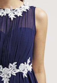 Námořnické šaty s plisé designem, průsvitným výstřihem a složitou bílou květinovou výšivkou podél límce a pasu.
