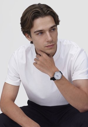 Mladenič z rjavimi lasmi, oblečen v belo majico in črne hlače, si spočije brado na roki, nosi uro s črnim usnjenim paščkom.
