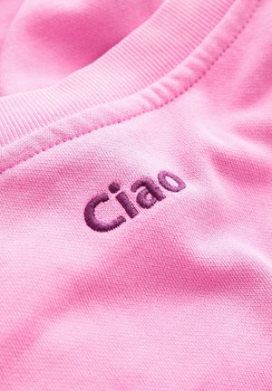 Roze stof met een gladde textuur, voorzien van het geborduurde woord "Ciao" in donkerpaars, nabij een geribde halslijn.