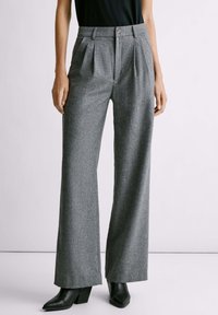 Pantalones grises ajustados con un diseño de pierna amplia, que presentan pliegues frontales, un cierre de botón y un acabado texturizado, combinados con botas de tacón negras.
