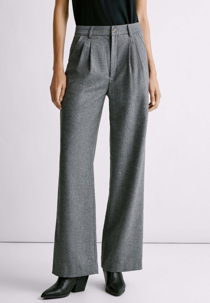 Pantalones grises ajustados con un diseño de pierna amplia, que presentan pliegues frontales, un cierre de botón y un acabado texturizado, combinados con botas de tacón negras.