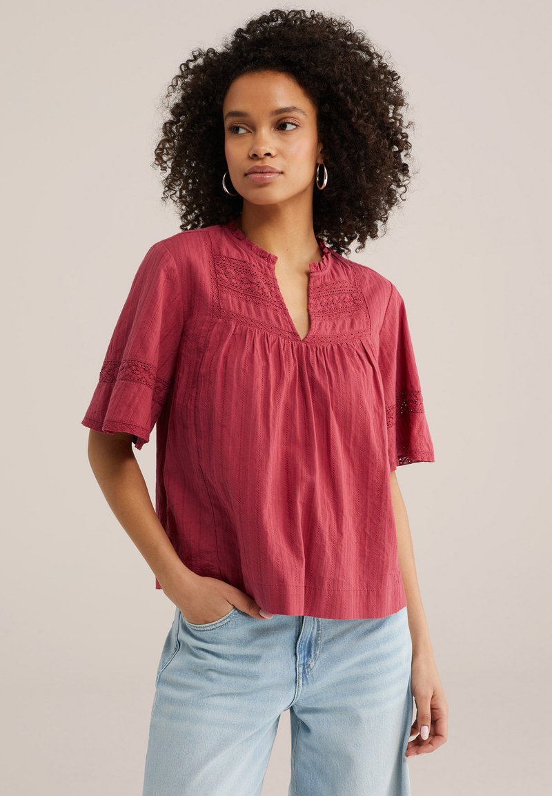 Femme aux cheveux bouclés portant des boucles d'oreilles anneau, un chemisier rouge à manches courtes avec des détails en dentelle, et un jean bleu clair, les mains dans les poches, regardant sur le côté.