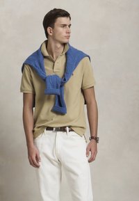 Jauns vīrietis, tērpts beigu polo krekls, baltas bikses, ar zilu džemperi, kas uzsiets plecos, skatās sānis pret tukšu fonu.