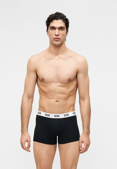 Homme musclé aux cheveux noirs courts portant un boxer noir avec une ceinture élastique blanche sur laquelle est inscrit "GCDS", sur un fond uni.