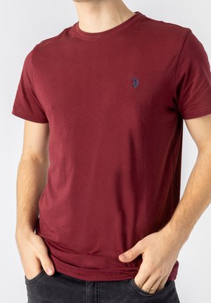 Mężczyzna noszący gładki bordowy t-shirt z krótkim rękawem, z małym granatowym haftowanym logo na piersi, ręce w kieszeniach czarnych dżinsów.