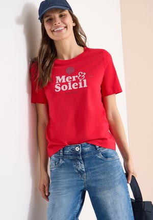 Femme souriante portant un t-shirt rouge "Mer & Soleil", un jean bleu et une casquette marine, tenant un sac noir et s'appuyant contre un mur.