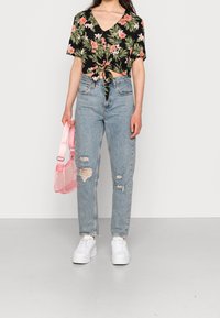 Bluse mit kurzen Ärmeln in Schwarz mit floralem Muster und geknotetem Vorderteil, kombiniert mit hochgeschnittenen, hellblauen, zerrissenen Jeans und weißen Plateauschuhen. Hält eine rosa Tasche.