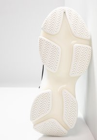 Semelle d'une basket avec une semelle extérieure en caoutchouc blanc texturé présentant des motifs en zigzag, une semelle intermédiaire beige lisse et une partie supérieure noire.