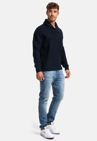 Granatowy sweter dziany z kołnierzem szalowym, noszony na tle zniszczonych jasnoniebieskich dżinsów i białych sneakersów, ukazujący swobodny styl.