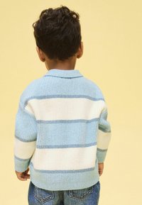 Vaaleansininen ja kermanvärinen raidallinen neuletakki, jossa on resoripinta; siinä on kauluksellinen pääntie ja pitkät hihat, yhdistetty farkkuihin.