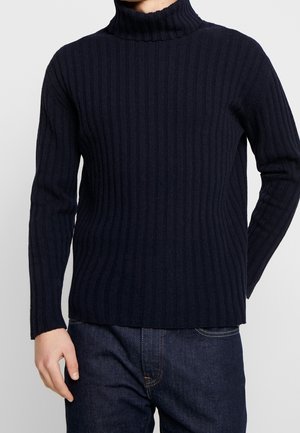 Mand iført en mørk navy ribbet rullekravetrøje og mørkeblå jeans mod en ensfarvet baggrund.