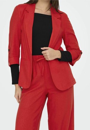 Frau trägt einen roten Blazer und passende rote Hose mit Kordelzug über einem schwarzen Langarmshirt, Hand hält den Revers des Blazers.
