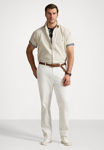 Collared beige shirt with short sleeves, layered over a striped top. Paired with white trousers and a braided brown belt. White sneakers.