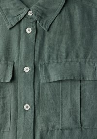 Camicia verde oliva con bottoni in lino, caratterizzata da un colletto strutturato, due tasche sul petto e una sottile texture. Bottoni bianchi offrono un contrasto.