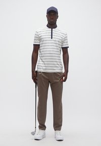 Άνδρας με ριγέ γκολφ πουκάμισο, καφέ παντελόνι, λευκά αθλητικά και ναυτικό καπέλο κρατώντας ένα γκολφ κλαμπ, στέκεται μπροστά σε απλό φόντο.