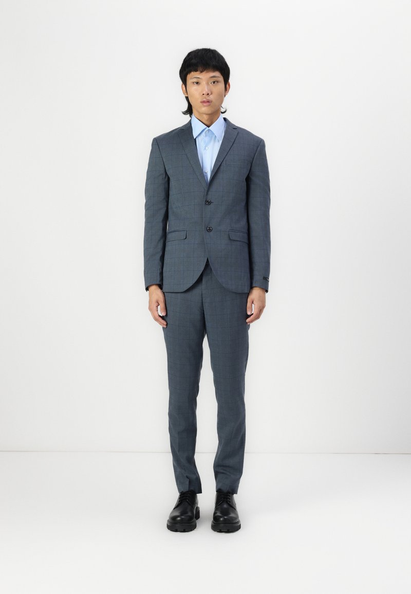 Abito grigio scuro a quadri con rever a punta, bottoni e tasche; abbinato a una camicia azzurra chiara e scarpe nere; design slim fit.