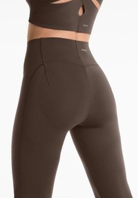 Leggings marron taille haute en matériau extensible, avec des détails de couture et une petite poche sur le côté. Découpes au niveau de la taille.