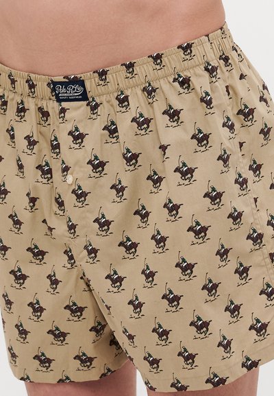 Beige bokserit, joissa on toistuva kuvio ratsastajista hevosilla. Valmistettu pehmeästä kankaasta, jossa on joustava vyötärö ja yksittäinen napitus.