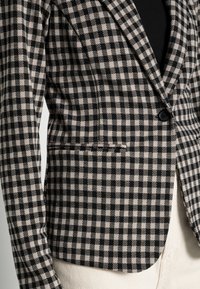 Blazer à carreaux noirs et beiges avec un revers cranté, une fermeture à un bouton et une poche latérale. Tissu texturé avec une coupe ajustée.