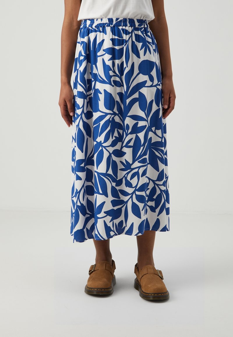 Nainen, jolla on päällään valkoinen hame sinivihreillä lehtikuosisilla ja ruskeat solkipohjaiset kengät, seisoo yksinkertaisella valkoisella taustalla.