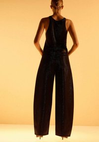 Femme aux cheveux blonds en chignon bas, portant un haut sans manches noir et un pantalon large, se tenant avec les mains sur les hanches devant un fond beige.