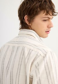 Camisa de color claro con rayas grises verticales, que presenta un cuello con solapa y una tela texturizada, vista desde la espalda.