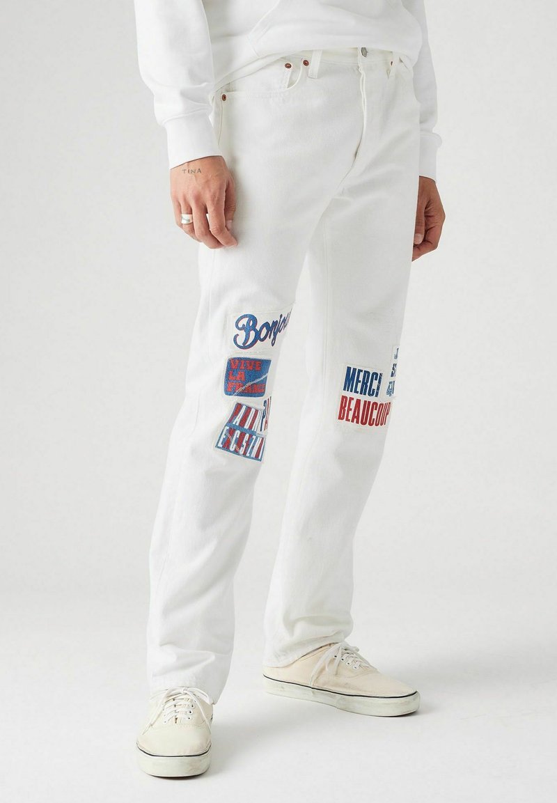 Onderlichaam gekleed in witte jeans met rode en blauwe Franse tekstpatches en witte sneakers op een lichtgrijze achtergrond.