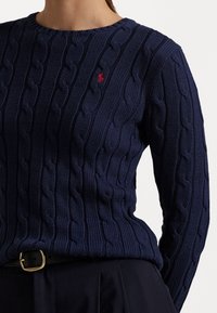 Žena v námořnickém modrém svetr s tváří v úpletu a malým červeným logem na hrudi, zasunutým do černých kalhot s černým páskem a zlatou sponou.