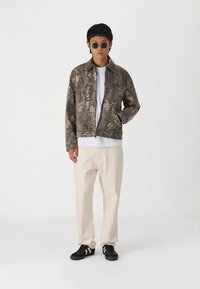 Veste zippée à motif camouflage dans des nuances de brun, portée par-dessus une chemise blanche, associée à un pantalon crème et des baskets noires.