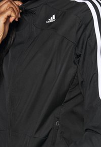 Chaqueta deportiva negra de tela ligera, con un bolsillo con cremallera, rayas blancas y un logo blanco de Adidas en el pecho.