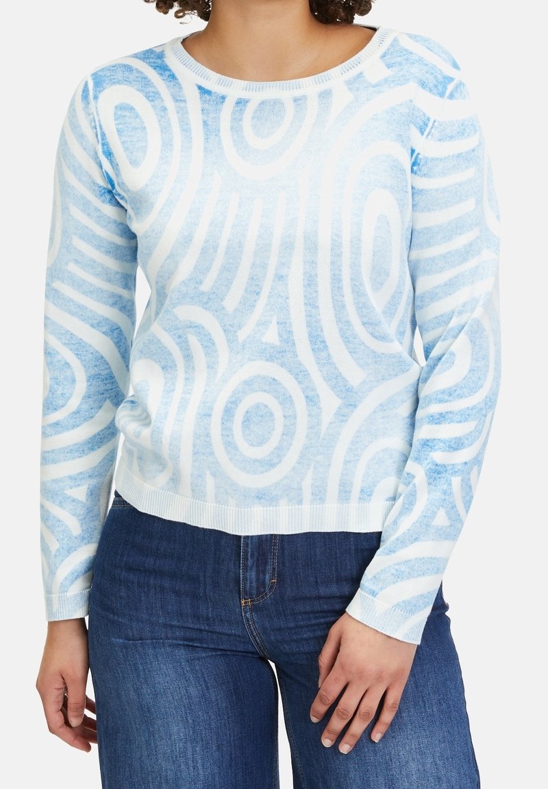 Femme portant un pull à manches longues à motifs bleu clair et blanc et un jean bleu foncé, debout avec une main détendue sur le côté.