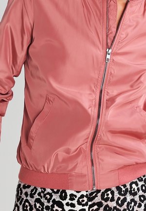 Veste bomber en satin rose pâle avec fermeture éclair frontale et poches latérales portée sur un pantalon imprimé léopard noir et blanc.
