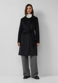 Cappotto avvolgente nero con collo a scialle, cintura annodata e tasche frontali. Tessuto morbido, design al ginocchio, abbinato a pantaloni larghi a coste grigi.
