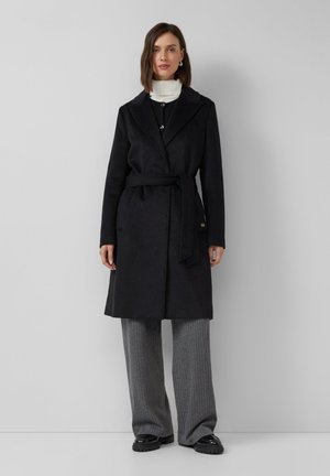 Manteau noir à col châle, ceinture nouée et poches avant. Tissu lisse, design longueur genoux, associé à un pantalon large en maille côtelée gris.